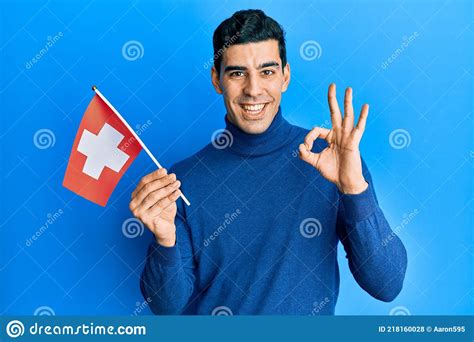 Handsome Hispanic Man Holding Switzerland Flag Doing Ok Sign With