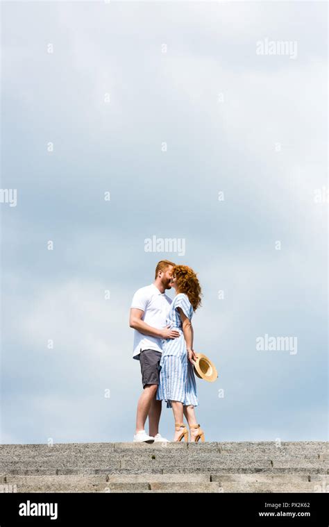 Low Angle View Of Redhead Man Embracing And Kissing Girlfriend On Stairs Against Cloudy Sky