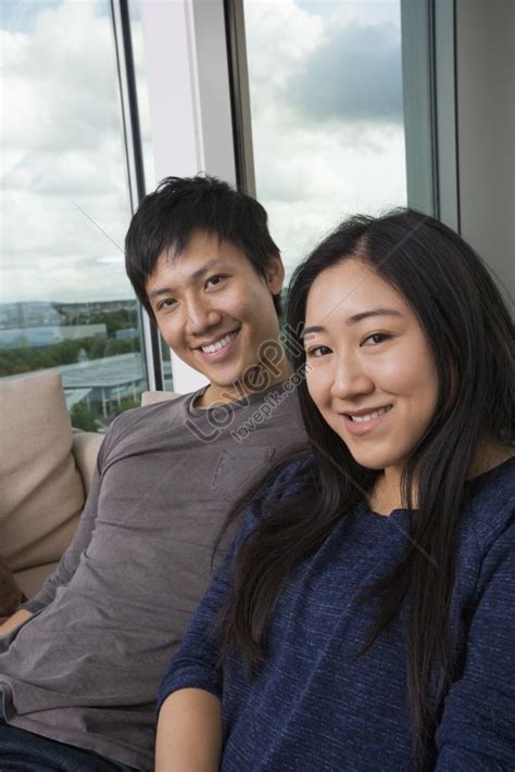 Portrait Of Asian Couple Sitting On Sofa At Home Smiling Happily