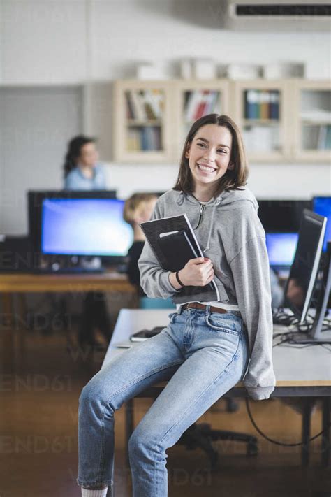 Cheerful High School Female Student Sitting With Books On Desk In