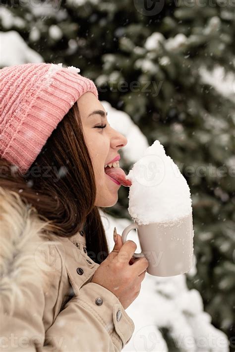 Woman In Warm Winter Clothes Standing By The Big Christmas Tree Outdoors And Licking Snow From