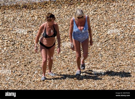 Two Older Ageing Women Walking On A Shingle Beach On A Hot Summers Day Wearing Swimming Costumes