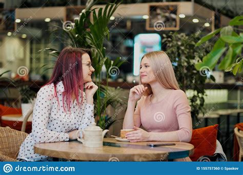 Two Young Beautiful Girls Sit In A Green Cafe At A Table