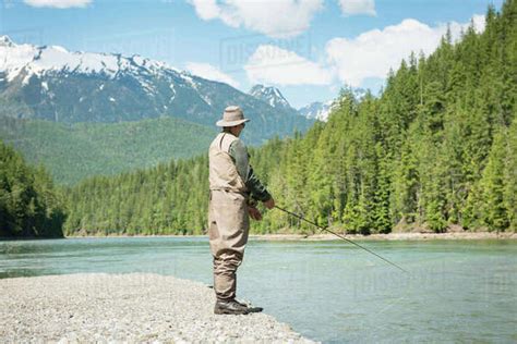 Full Length Of Man Fishing In River While Standing On Riverbank Against Mountain Stock Photo