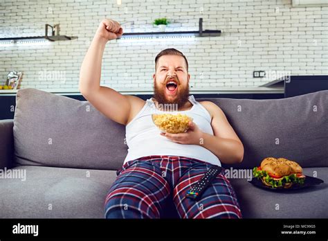 L homme drôle épais en pyjama de manger un hamburger assis sur le