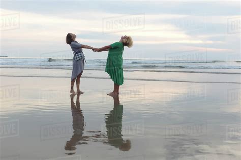 Full Length Of Happy Female Friends Holding Hands While Standing On Shore At Beach During Sunset