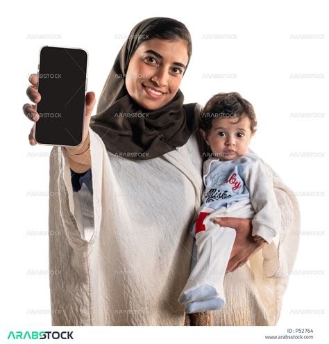 بورتريه لأم عربية خليجية سعودية تحمل طفلها الرضيع بسعادة وحنان وبيدها الهاتف المحمول، تعرض شاشة