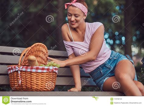 Happy Beautiful Redhead Female Wearing Casual Clothes Sitting With A Picnic Basket On A Bench In