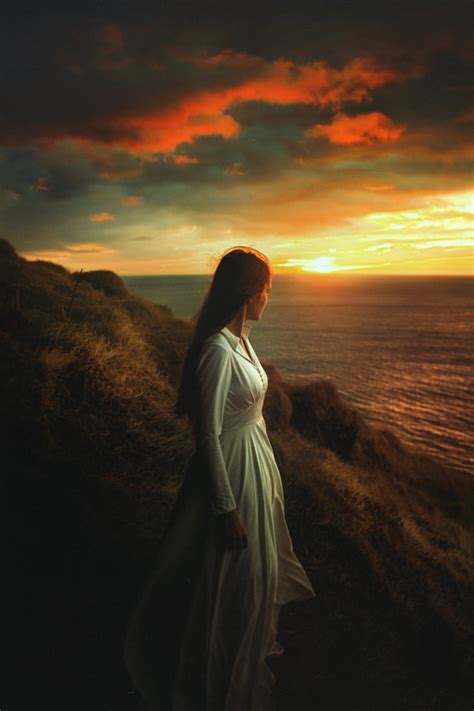 Фото Девушка в длинном белом платье стоит на обрыве у моря на фоне заката фотограф TJ Drysdale