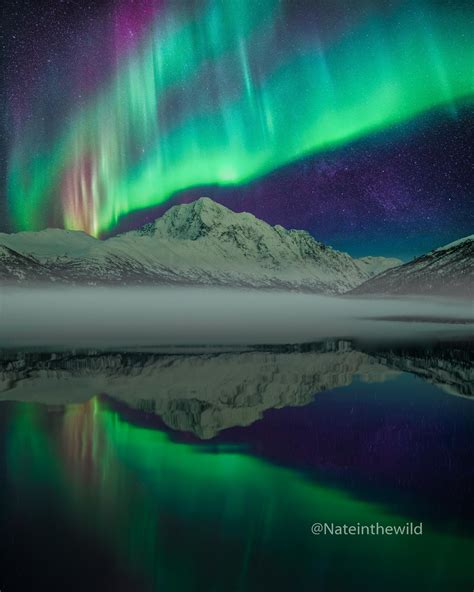 The Aurora Bore Is Reflected In Water With Mountains And Snow Capped
