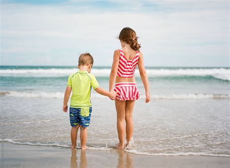 Big Sister Holding Little Brothers Hand While Walking Towards The Ocean By Stocksy
