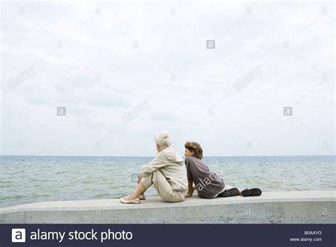Grandmother And Grandson Sitting Together By The Sea Looking At View