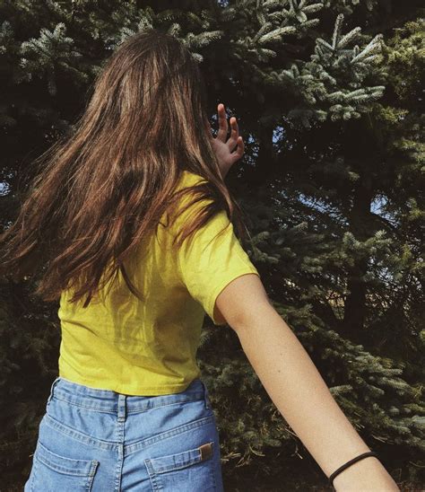 a woman with long brown hair wearing a yellow shirt and blue jean shorts standing in front of trees