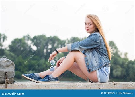 Young Beautiful Blonde Woman In Short Shorts And Jeans Sitting On The Waterfront In Summer And