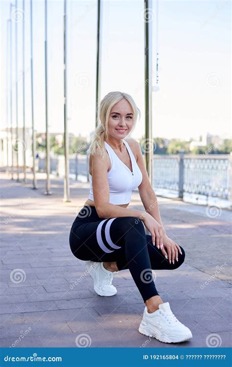 Young Fit Blond Woman Wearing White Top And Black Leggings Squatting Down Posing By City Lake