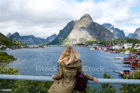 若い女性が立ってロフォーテン諸島の漁村を見ていますレーヌフィヨルドの海岸にある赤いスカンジナビアの家ノルウェーの観光名所 1人のストック