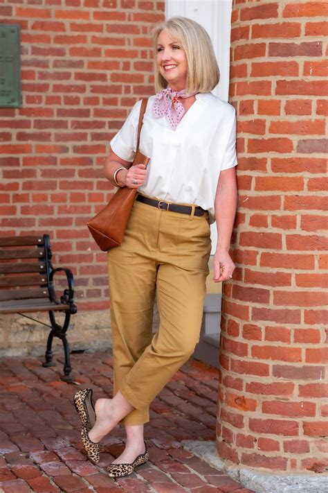 A Woman Standing Next To A Brick Wall Wearing Brown Pants And A White