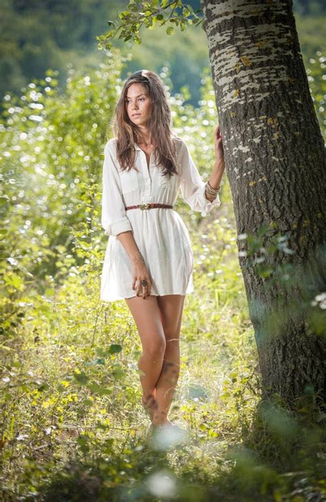attraktiv ung kvinna i den vita korta klänningen som poserar nära ett träd i en solig sommardag