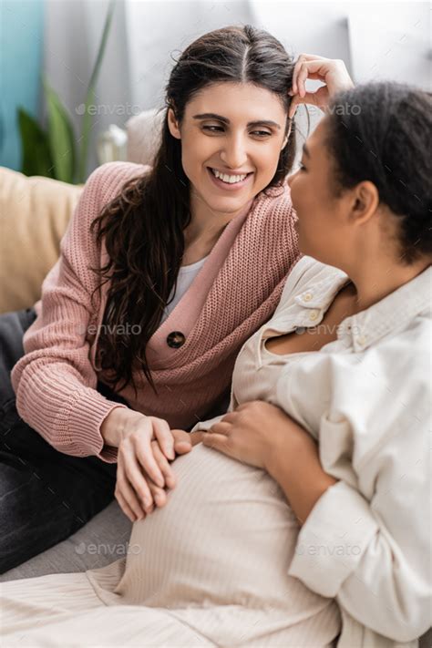 Happy Lesbian Woman Touching Tummy Of Pregnant Multiracial Partner And Sitting On Couch Stock