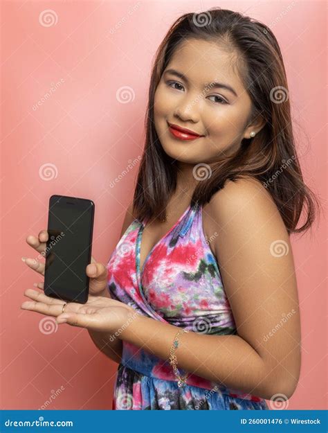 beautiful filipina in a colorful dress smiling and marketing a phone on a pink background stock