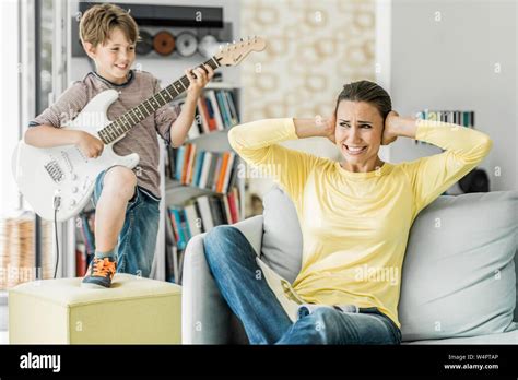 La madre se sienta en el sofá en el salón y cubre sus orejas hijo toca la guitarra eléctrica