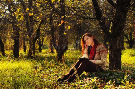 Young Woman With Long Red Hair Reading Under The Tree Stock Photo