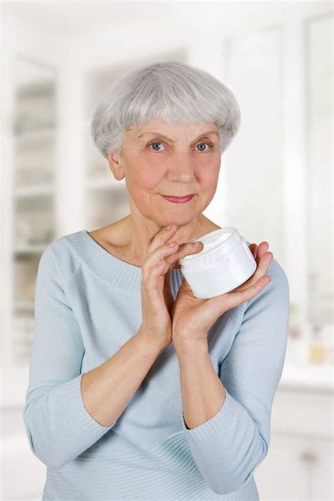 Charming Elderly Woman Applying Cosmetic Cream On Her Face For Facial Skin Care In Bathroom At