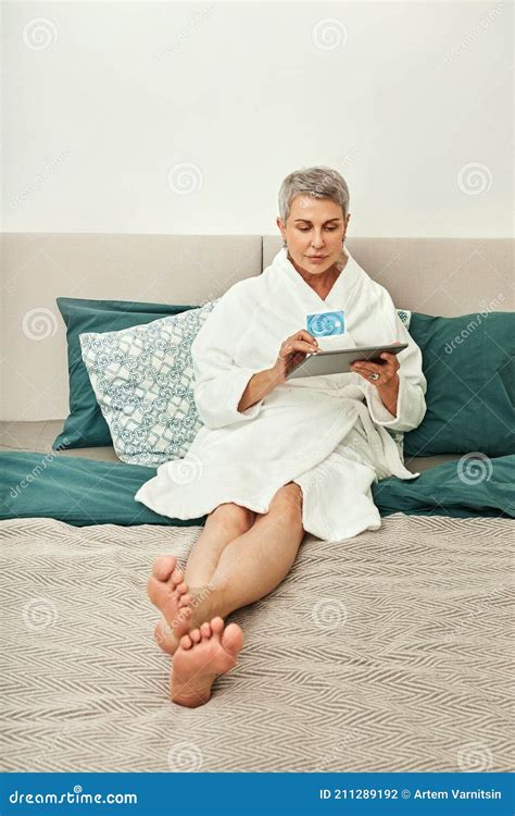 Mature Woman In Bathrobe Lying On A Bed Holding A Digital Tablet And