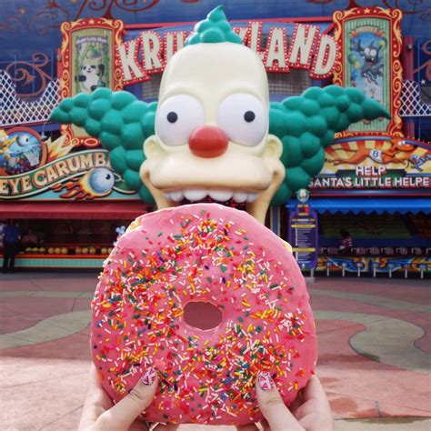 A Person Holding A Donut With Sprinkles In Front Of A Carnival