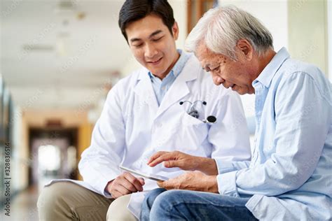 Young Asian Doctor Talking To Senior Man In Hospital Hallway ภาพถ่าย