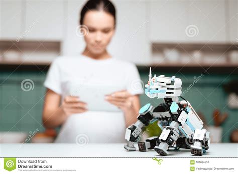 the girl is standing in the kitchen and holding a tablet a small rhinoceros robot sits next to