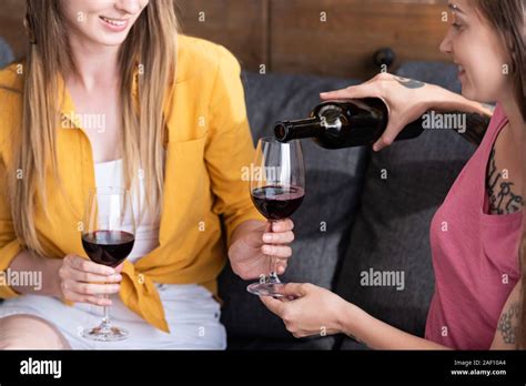 partial view of two lesbians holding wine glasses and pouring wine while sitting on sofa in