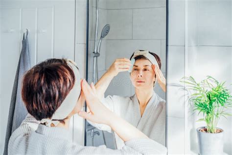 Woman In Bathrobe And Hair Band Looking In The Mirror And Making Face Massage With Gua Sha