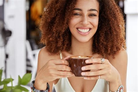 Photo Recadrée De Belle Femme Afro Américaine Joyeuse Tient Une Tasse De Café Déjeune Dans Une