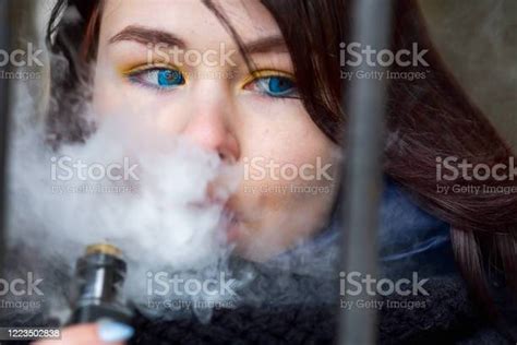 Vape 십대 푸른 눈과 푸른 재킷과 젊은 예쁜 흰색 백인 갈색 머리 소녀는 봄에 거리에서 오래된 금속 게이트 근처 전자 담배를 흡연 나쁜 습관 전자담배에 대한 스톡 사진