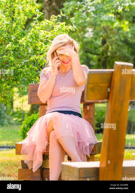 blonde woman sitting in park peeking at camera smiling while talking using on smartphone phone