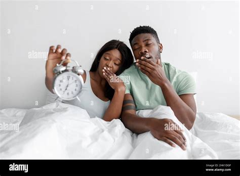 tired sad sleepy millennial african american couple sit on white bed in bedroom yawn and show