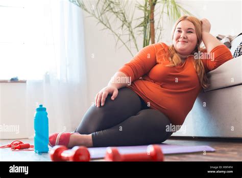 Chubby Woman Sport At Home Sitting On Yoga Mat Leaning On Sofa Smiling Relaxed Looking Aside