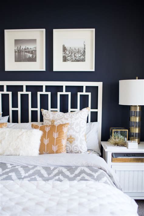 White and navy blue bedroom features an accent wall painted navy blue lined with a white curved nailhead headboard on bed dressed in soft white bedding flanked by white campaign nightstands placed below windows alongside a pair of leopard x benches placed at the foot of the bed. A Look Inside A Blogger's Navy and Mustard Bedroom - My ...