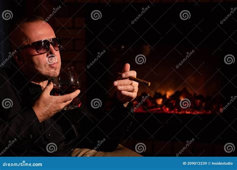 A Mature Man With Glasses Drinks Brandy And Smokes A Cigar While Sitting In An Armchair By The