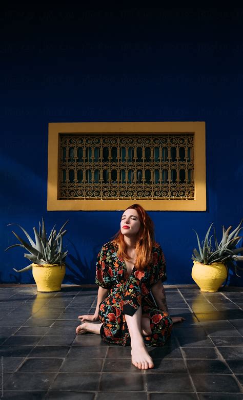 Portrait Of A Ginger Woman Sitting In Front Of A Blue And Yellow Facade Between Two Plant Pots