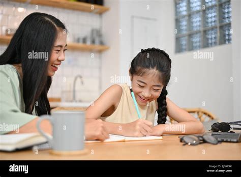 happy and kind asian mom teaching her daughter doing homework at dining table in modern home