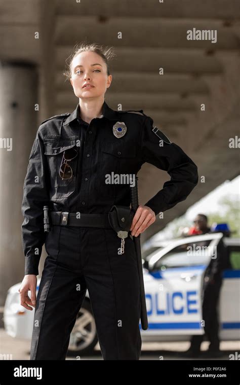 Attractive Female Police Officer In Uniform Looking At Camera With Blurred Partner Near Car On