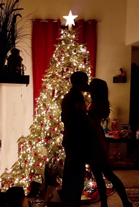 A Man And Woman Standing In Front Of A Christmas Tree With Lights On It