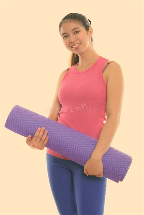 Studio Shot Of Young Happy Asian Woman Smiling While Holding Yoga Mat