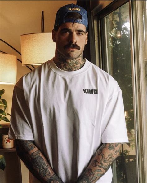 A Tattooed Man Standing In Front Of A Window Wearing A White Shirt And Blue Hat