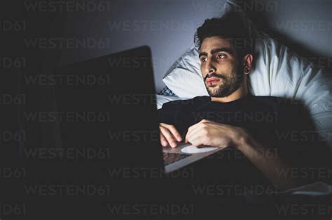 Bearded Young Man Lying In Bed At Night Looking At Laptop Stock Photo