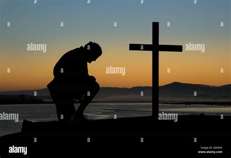 A Man Kneeling In Prayer By A Cross On A Hill At Dawn Of A New Day