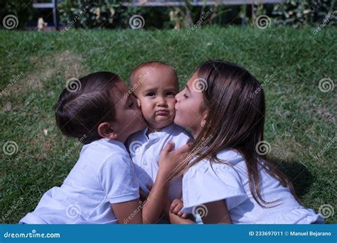 Brothers Sitting On The Grass In A Park Kissing Their Little Brother