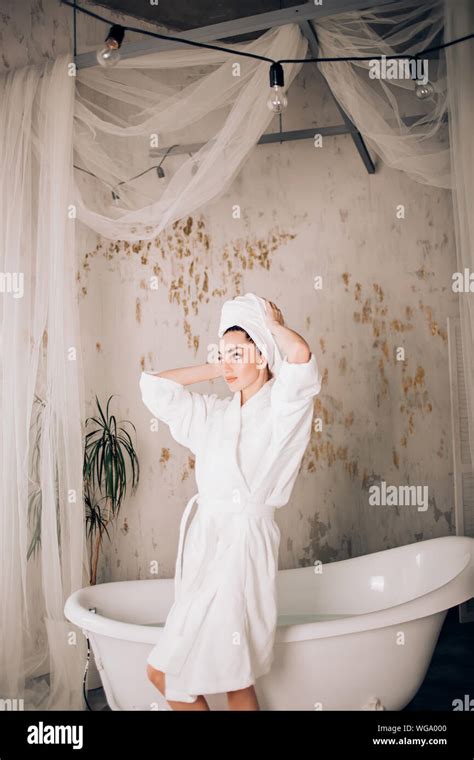 Young Beautiful Woman Wearing White Bathrobe Standing Near Bathtub Touching Towel On Her Head